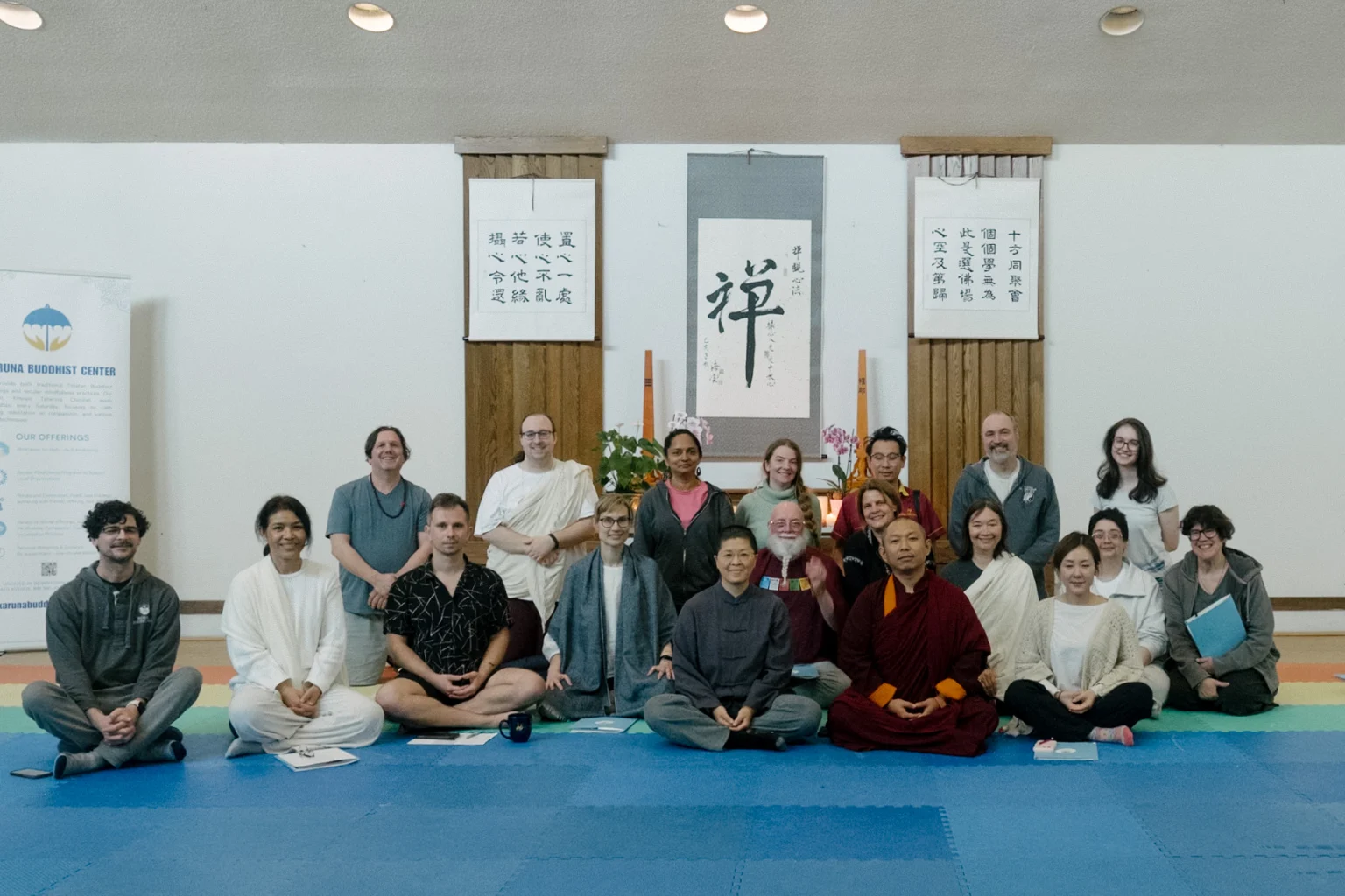 A large group of practitioners, including a lama in maroon robes, seated and standing on blue mats in a zendo hung with Chinese calligraphy scrolls