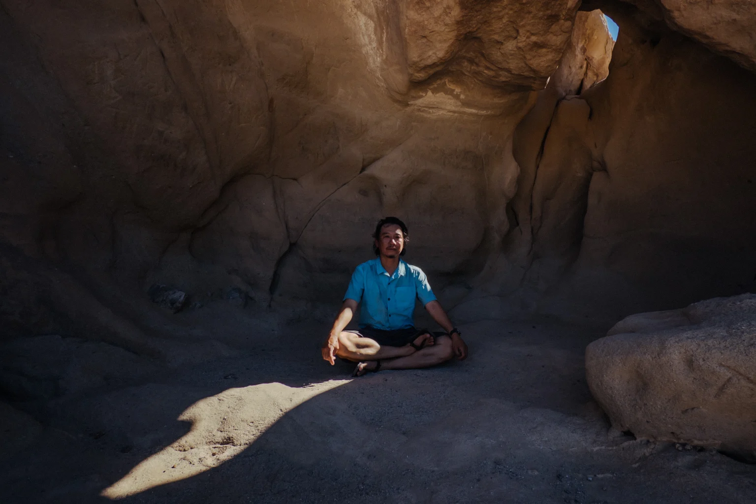 A man in a blue shirt sits cross-legged in meditation inside a wind-carved sandstone cave, a shaft of light on the floor