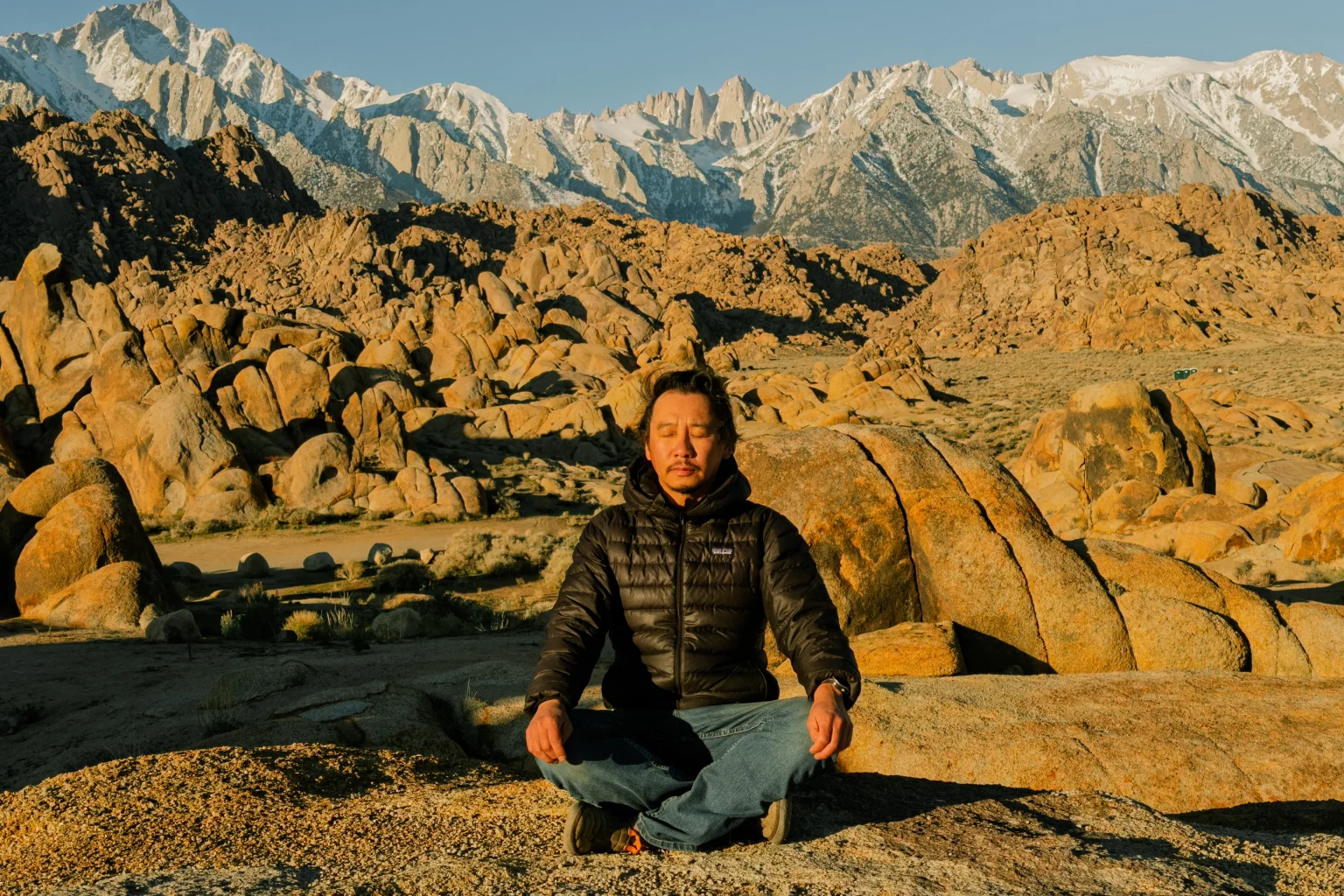 A man in a black puffer jacket meditates cross-legged on a granite boulder at sunrise, rounded rock formations and snow-covered peaks behind