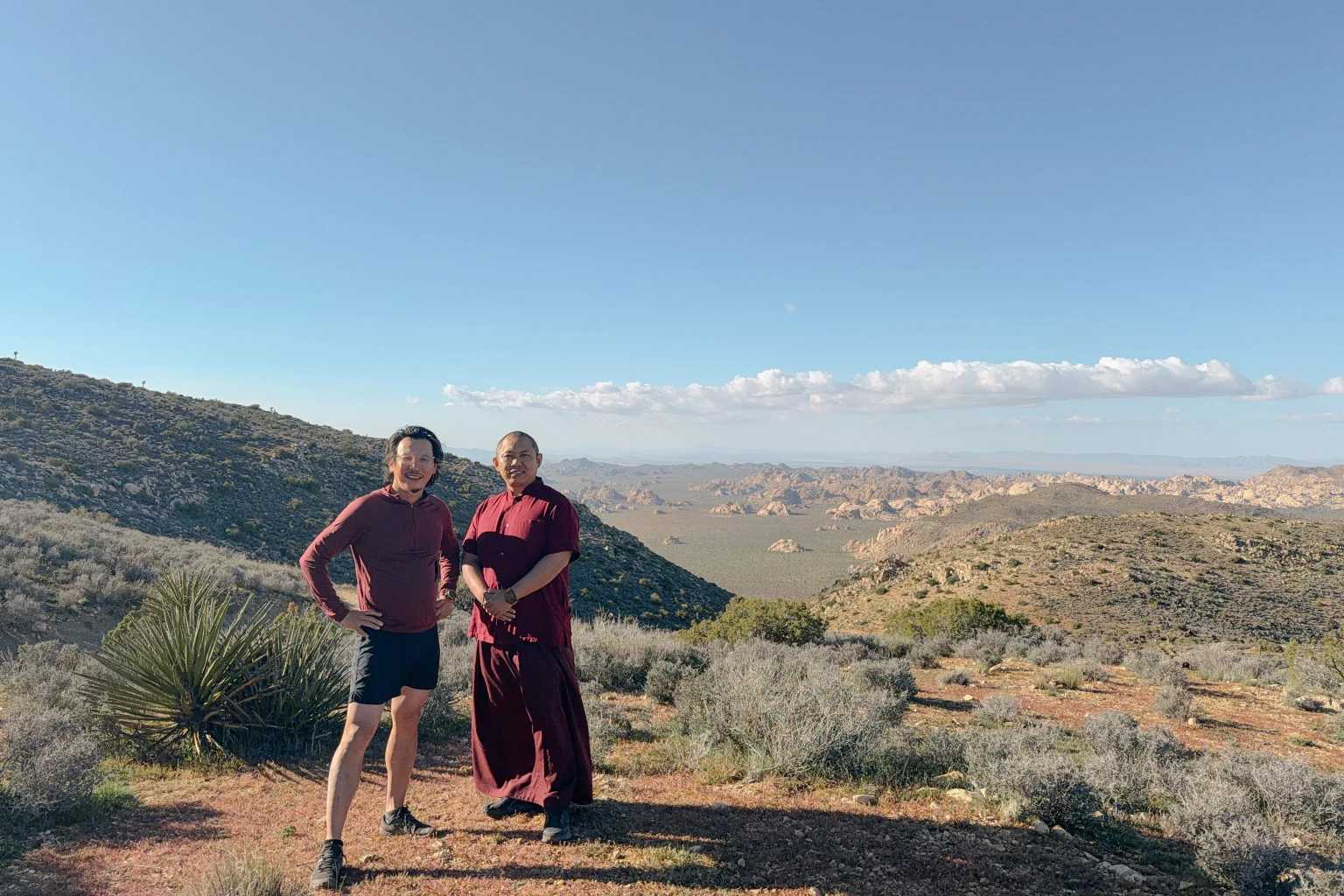 Two people, one in maroon monastic robes, stand on a desert trail with yucca and a wide valley of rocky hills behind