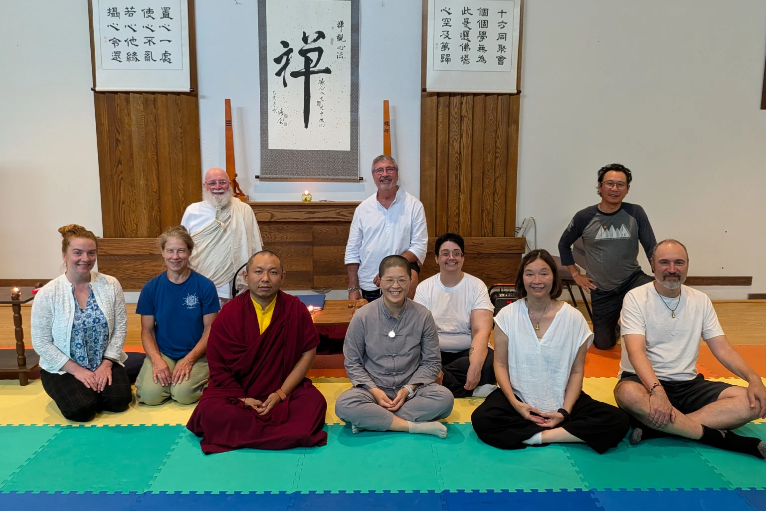 A small group, including a lama in maroon robes, sits together on colorful mats in front of an altar and calligraphy scrolls